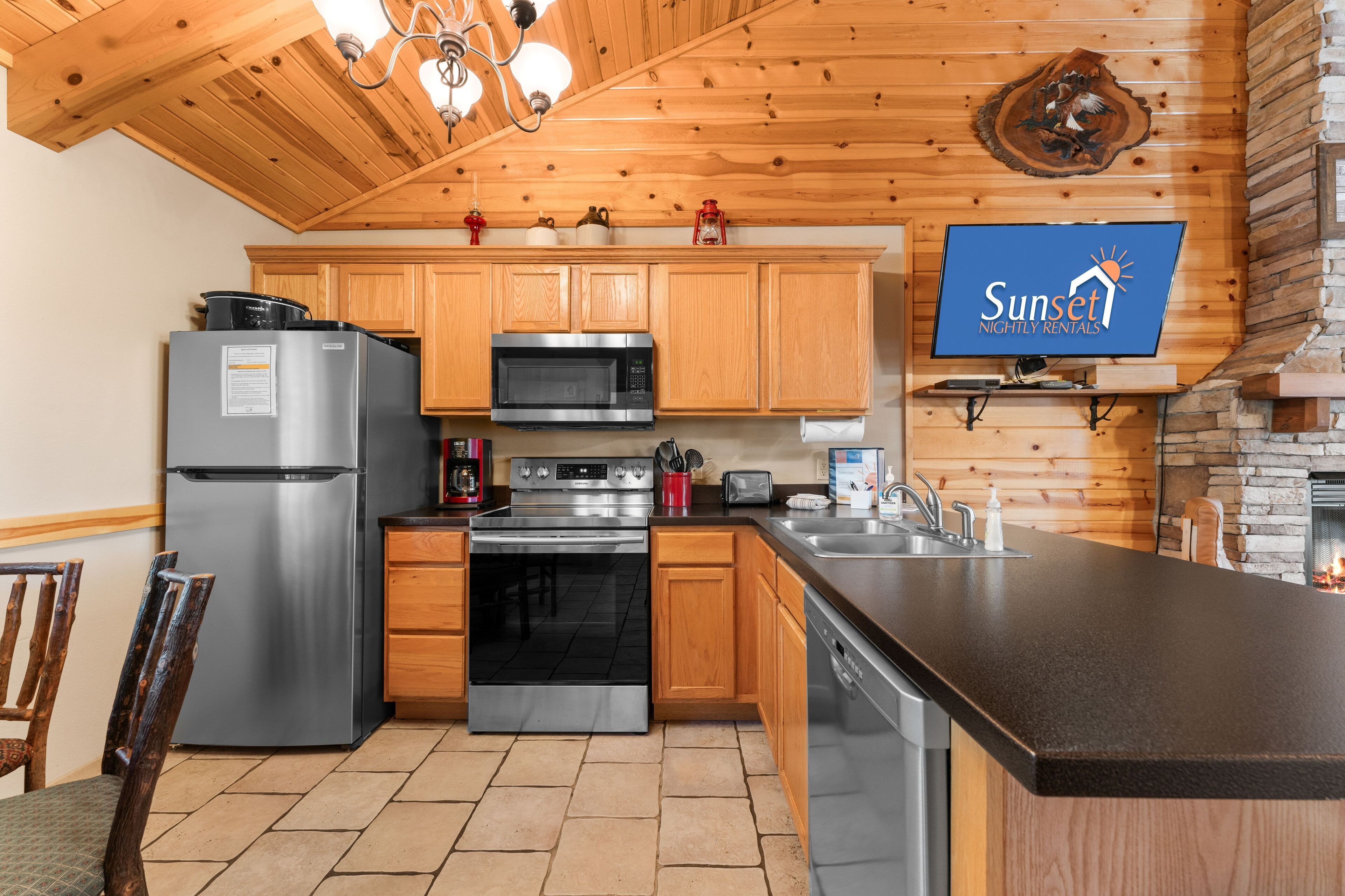 A modern kitchen with wooden cabinets, stainless steel appliances, a flat-screen TV, and a stone fireplace.