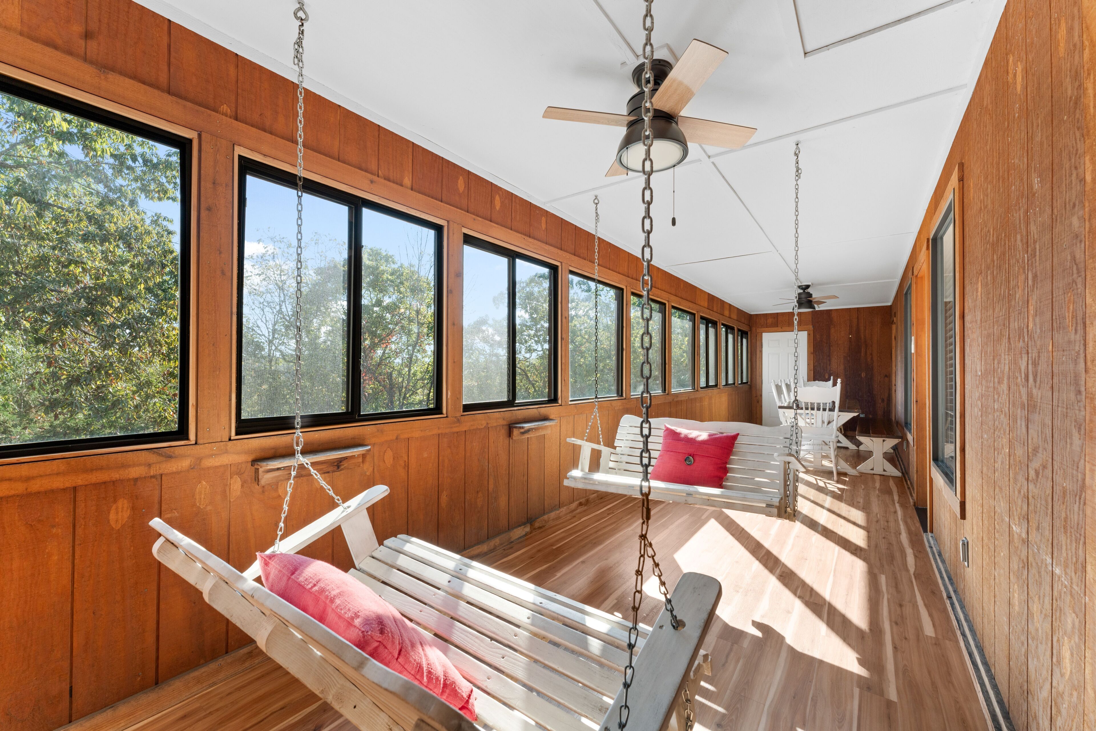 A cabin with large windows, wooden walls, and a ceiling fan. There are two swing seats with cushions.