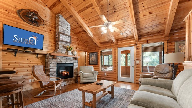 A log cabin interior with a fireplace, wooden ceiling, and a television displaying 'Sunset Nightly Rentals'. - Ozarks Oasis 4 Bedroom Cabin by RedAwning (Branson)