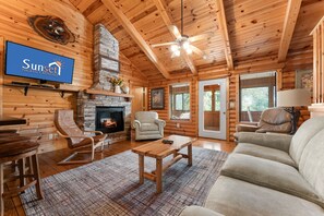 L’intérieur d’une cabane en rondins, avec une cheminée, un plafond en bois et un téléviseur diffusant une émission sur les locations de vacances au coucher du soleil.