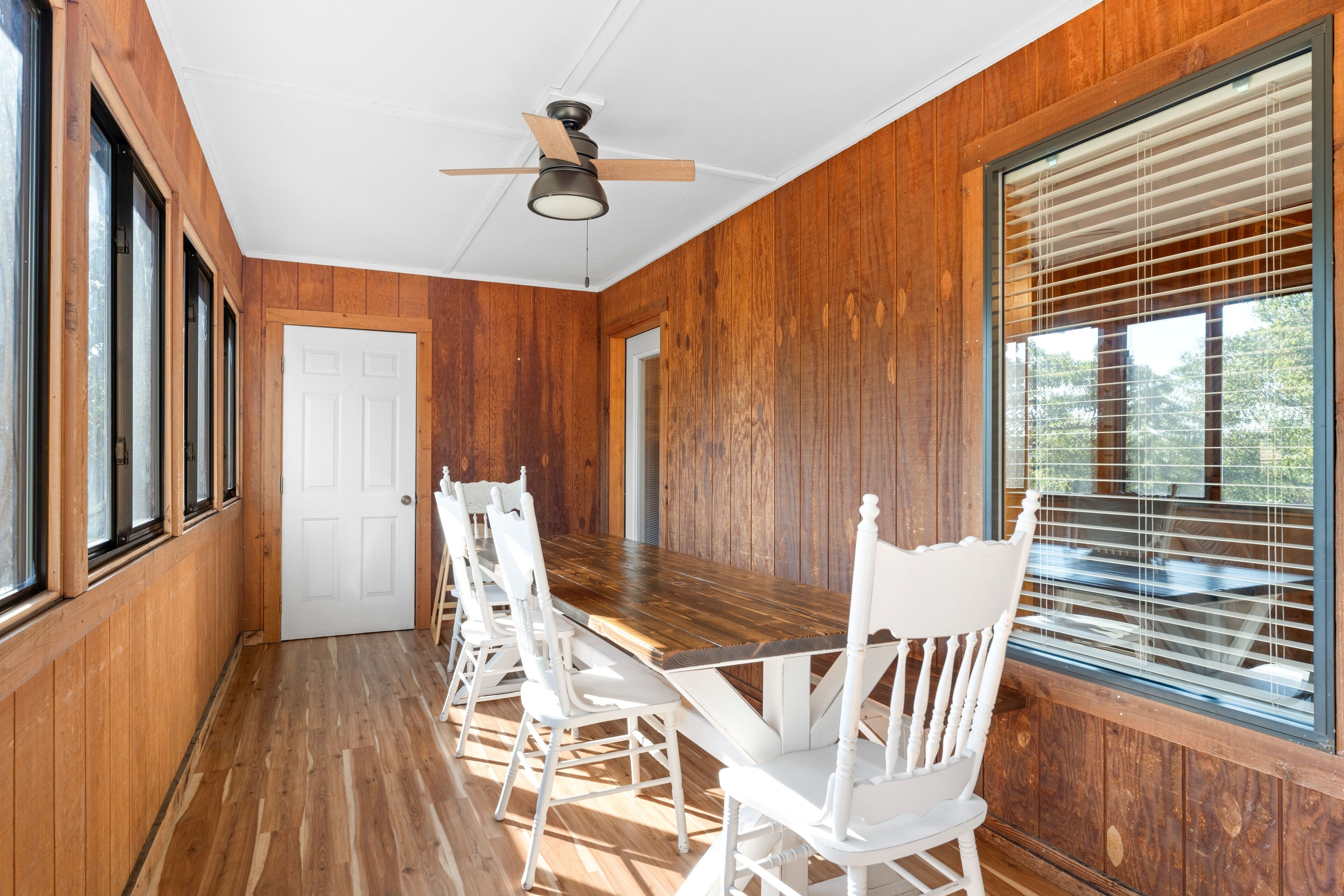 A wooden-paneled room with a large dining table, white chairs, and a ceiling fan.