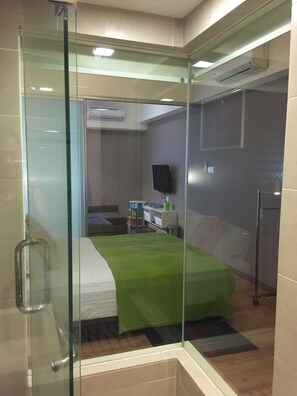 A modern hotel room with a glass shower enclosure, a bed with a green bedspread, a desk with a computer, and a flat-screen TV mounted on the wall.