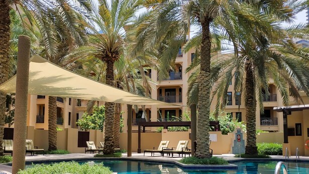 A pool area with lounge chairs and umbrellas, surrounded by palm trees and a building with balconies. - Maison Privee - Luxury Living Next to Dubai Mall & Burj Khalifa (Dubai)