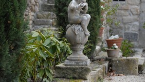 Une statue de pierre représentant une figure assise, placée dans un jardin, avec des plantes en pot et un mur de pierre en arrière-plan.