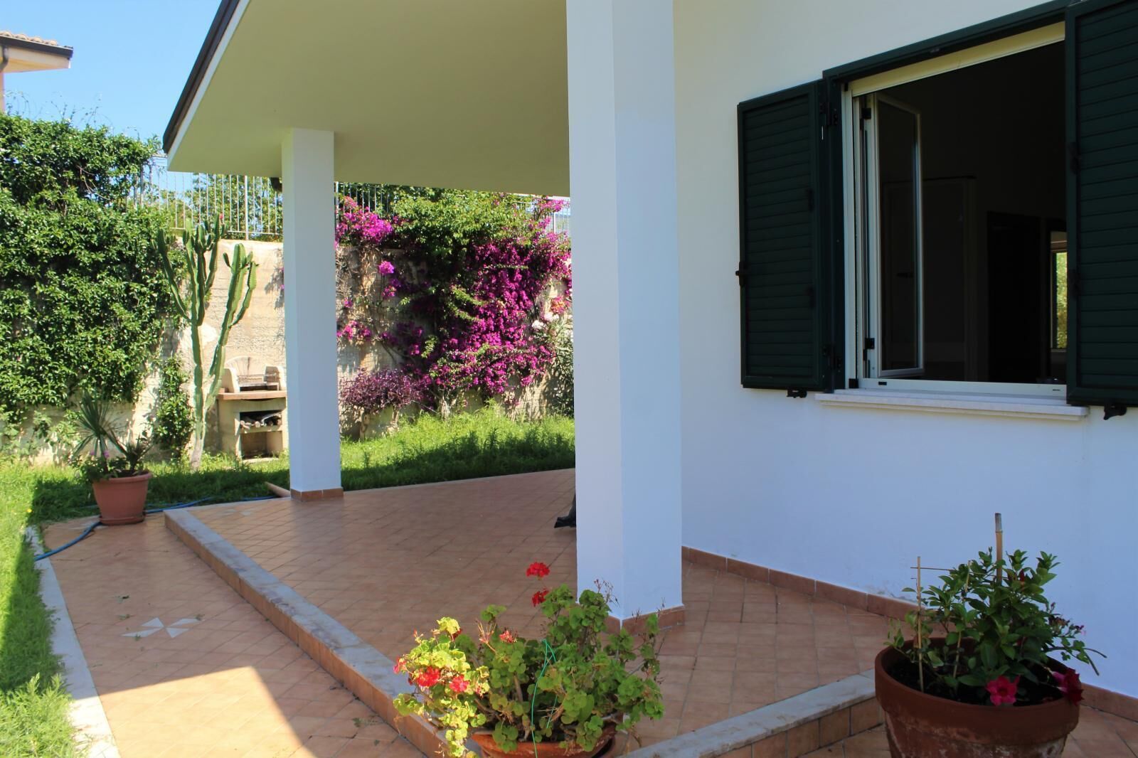 Un patio techado con suelo de baldosas, plantas en macetas y vistas a un jardín lleno de flores moradas.
