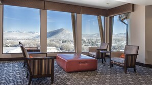 A hotel room with large windows offering a mountain view, a patterned carpet, and a brown ottoman.