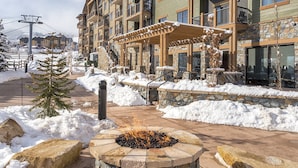 A snow-covered outdoor area with a stone fire pit, a wooden pergola, and a building with balconies and large windows.