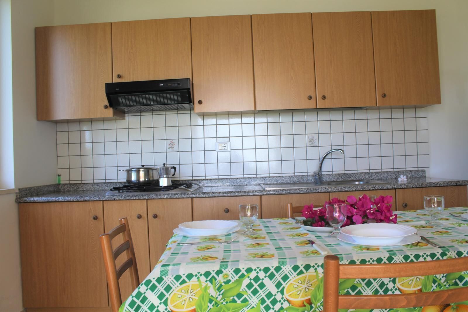 Eine Küche mit Holdekor-Schränken, gefliestem Fliesenspiegel, einem Herd und einem Tisch, der mit Geschirr und einer Blumenvase gedeckt ist.