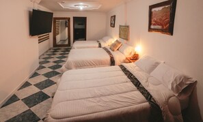 Habitación de hotel con tres camas alineadas, suelo a cuadros, televisor de pantalla plana en la pared y cuadros en las paredes.