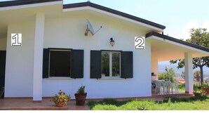 A two-story house with a covered patio, two windows with shutters, a satellite dish, and a view of the surrounding landscape. - Villa Faro with panoramic garden and barbecue. 350m from the sea (RICADI)