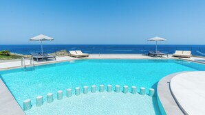 Une piscine d’un bleu éclatant, bordée de chaises longues blanches et d’ombrelles, entourée d’un mur de pierre et offrant une vue sur l’océan.