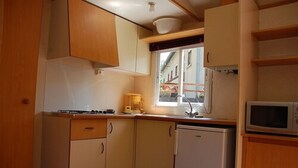 A compact kitchen with wooden cabinets, a microwave, a small refrigerator, and a window providing natural light. - Camping Les Airelles (Saint-Cirgues-en-Montagne)