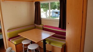 A compact kitchenette with a wooden table and two stools, a window with curtains, and a bench with colorful cushions. - Camping Les Airelles (Saint-Cirgues-en-Montagne)
