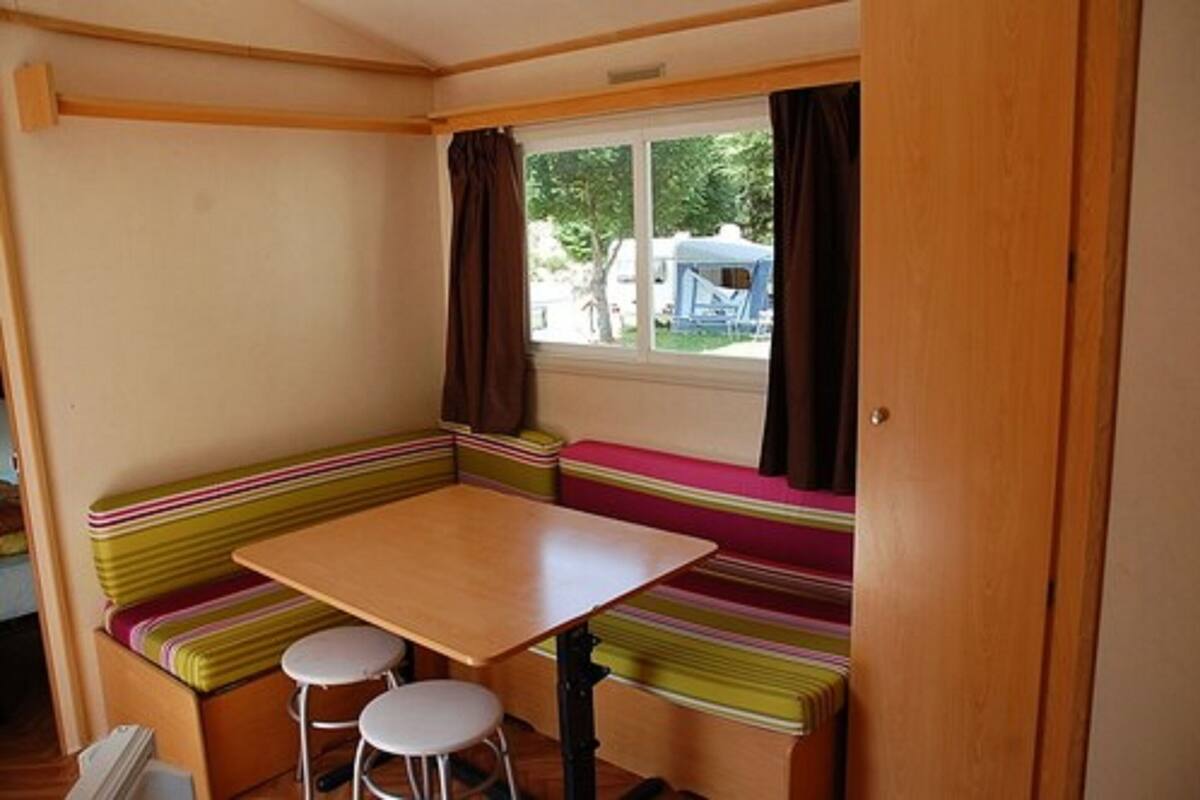Eine kleine Küchenzeile mit Holztisch und zwei Hocker, einem Fenster mit Vorhängen und einer Bank mit bunten Kissen.