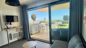 A modern living room with a sofa, a small table, a TV, and a large sliding glass door leading to an outdoor area with a view of the sea. - Apartment for 4 with terrace, garden, pool and beach access - Pets allowed (La Tranche-sur-Mer)