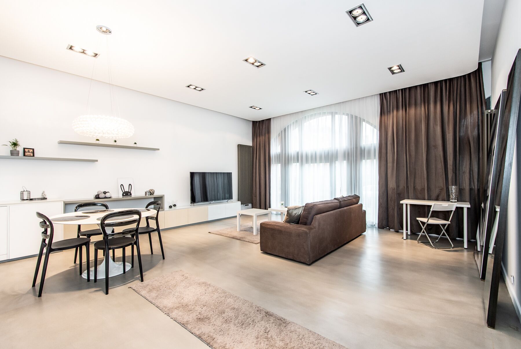 Ein modernes Wohnzimmer mit Sofa, einem Esstisch mit Stühlen, einem Fernseher und einem großen Fenster mit Vorhängen.