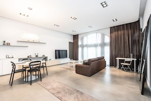 Uma sala de estar moderna com sofá, mesa de jantar com cadeiras, televisão e uma janela grande com cortinas.