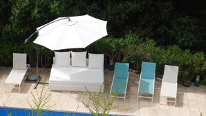 Un área junto a la piscina con un sofá blanco para exteriores, dos sillones reclinables y una sombrilla grande.
