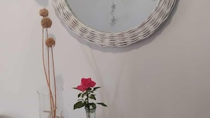Un espejo redondo con un marco tejido sobre una pared blanca, una mesita con un florero con una sola rosa, y un florero de vidrio con flores secas.