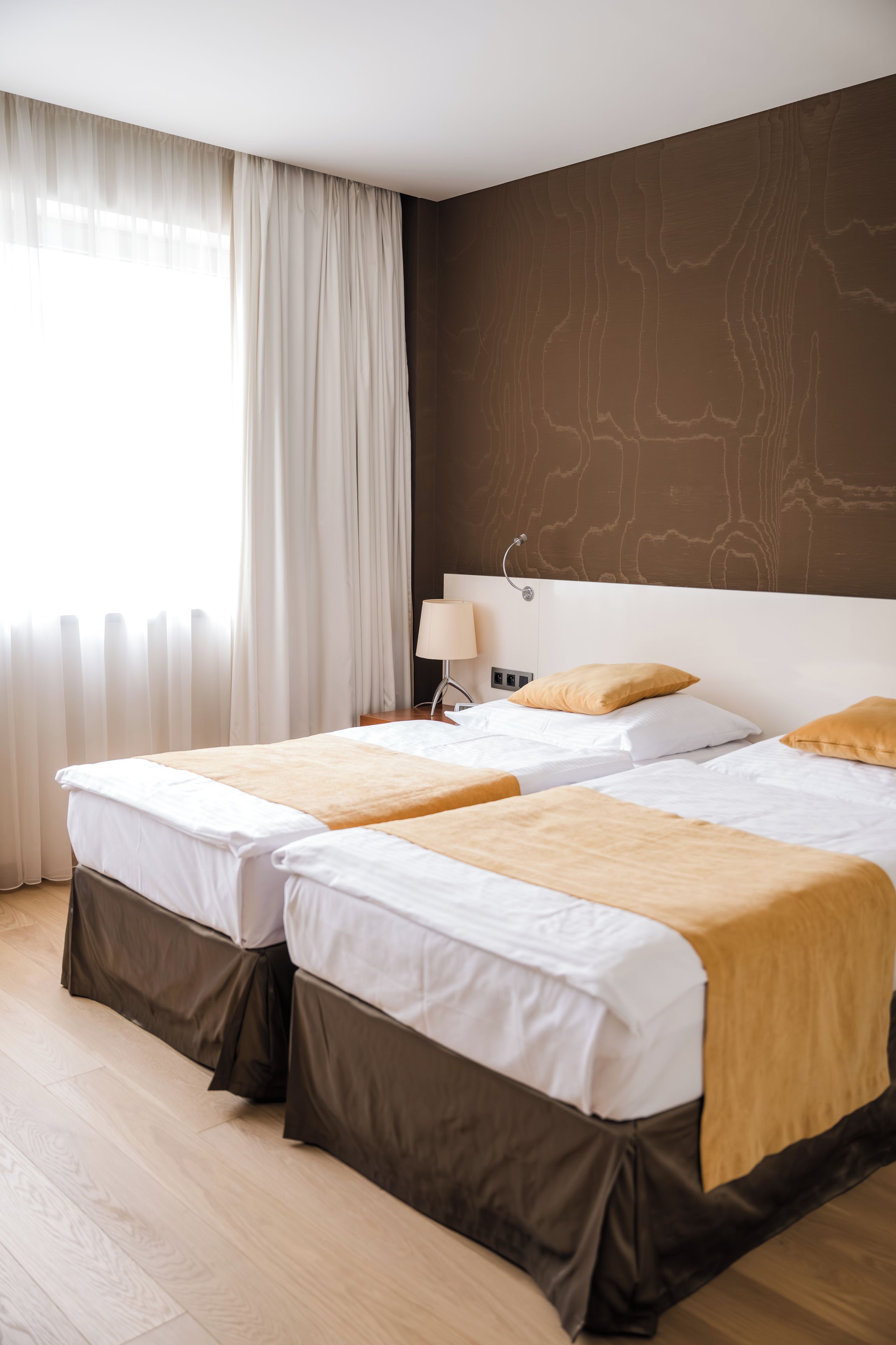 Ein Hotelzimmer mit zwei Betten, einem großen Fenster mit Vorhängen, einem Nachttisch mit Lampe und einer Wand mit Holzoptik.