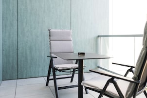 Un balcón con una silla y una mesita pequeña, piso de baldosas y una pared azul claro.