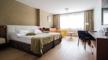 Habitación de hotel con cama, escritorio, silla, mesita, planta y ventana con cortinas.