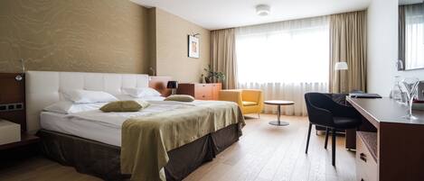 A hotel room with a bed, a desk, a chair, a small table, a plant, and a window with curtains.