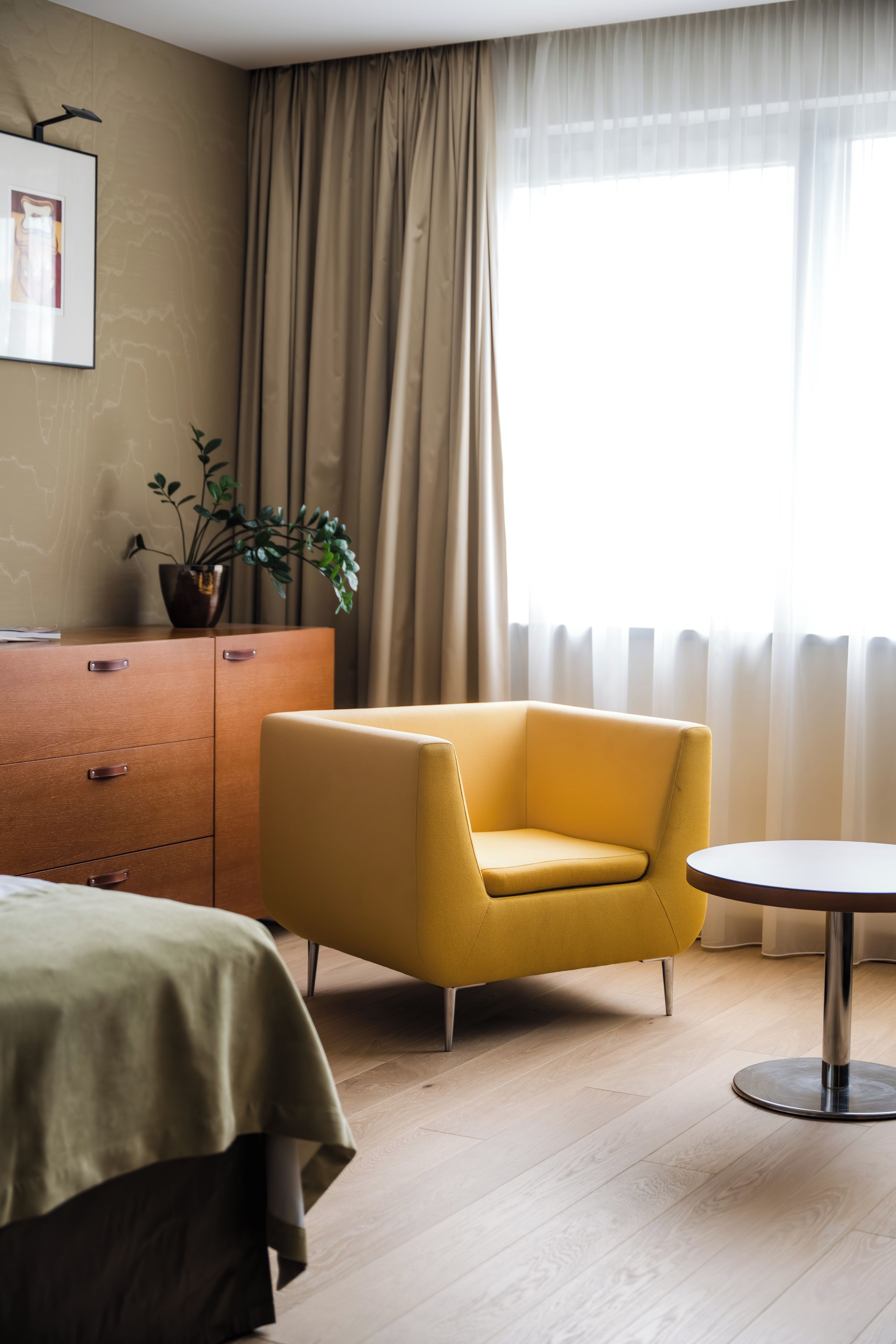 Una habitación de hotel con una cama, un sillón amarillo, una mesita, un aparador, una planta en maceta y una ventana con cortinas.