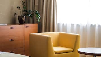 Una habitación de hotel con una cama, un sillón amarillo, una mesita, un aparador, una planta en maceta y una ventana con cortinas.