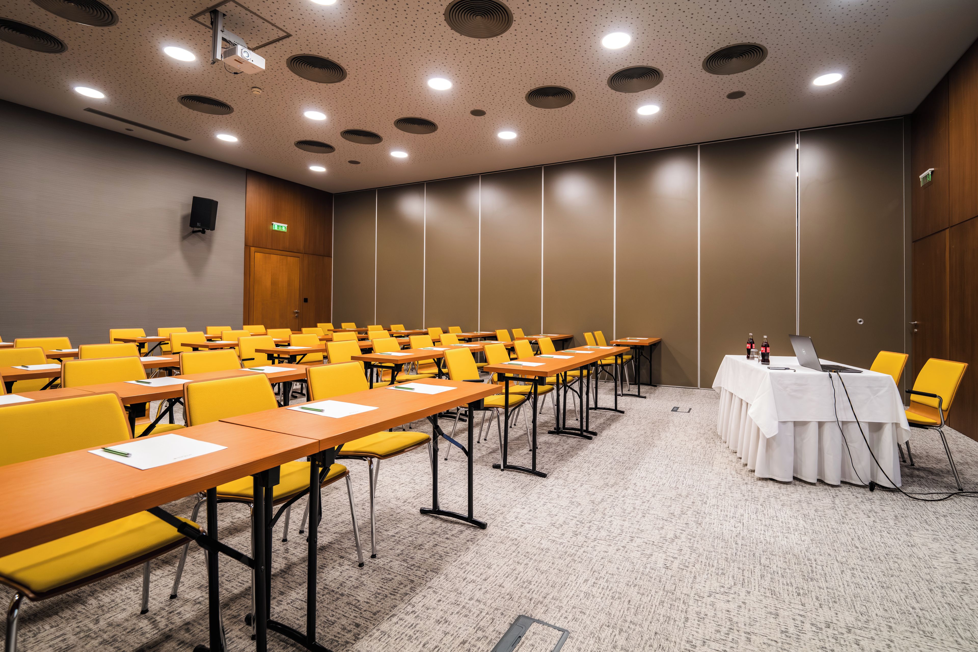 Une salle de conférence avec des rangées de chaises jaunes, une table blanche sur laquelle se trouvent un ordinateur portable et des bouteilles, et un projecteur fixé au plafond.