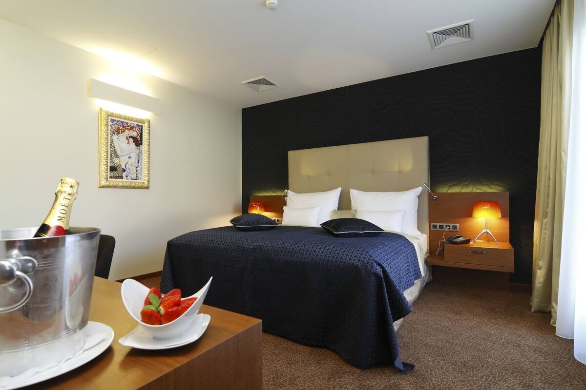 A hotel room with a large bed, a bedside table with a bottle of champagne, a bowl of strawberries, and a nightstand with a lamp.