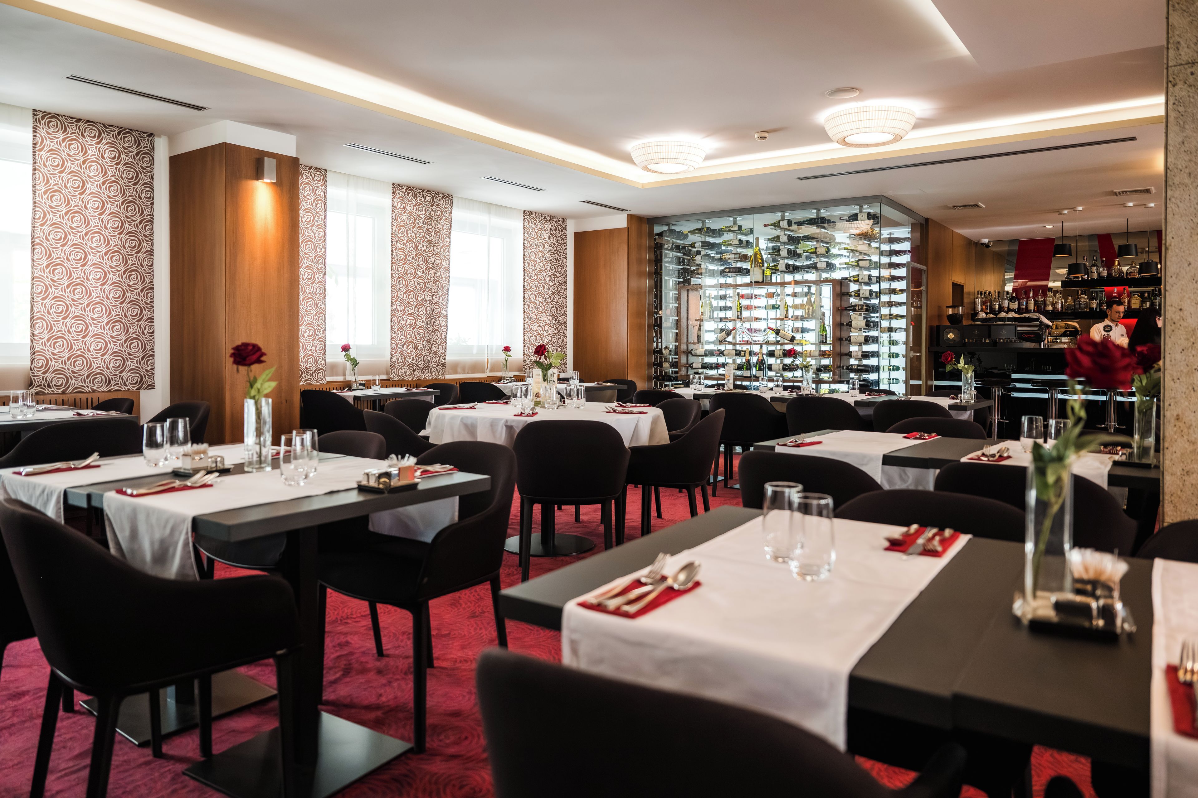 Un restaurant avec de nombreuses tables, un sol recouvert d’un tapis rouge et un présentoir à vin en arrière-plan.