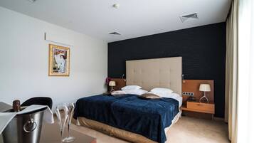 Quarto de hotel com cama, mesas de cabeceira, escrivaninha, luminária, cadeira e um quadro na parede.