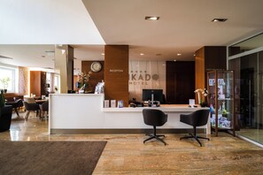 Zona de recepción de un hotel, con mostrador de recepción, dos sillas negras, un reloj y un cartel con el nombre del hotel y la recepción.