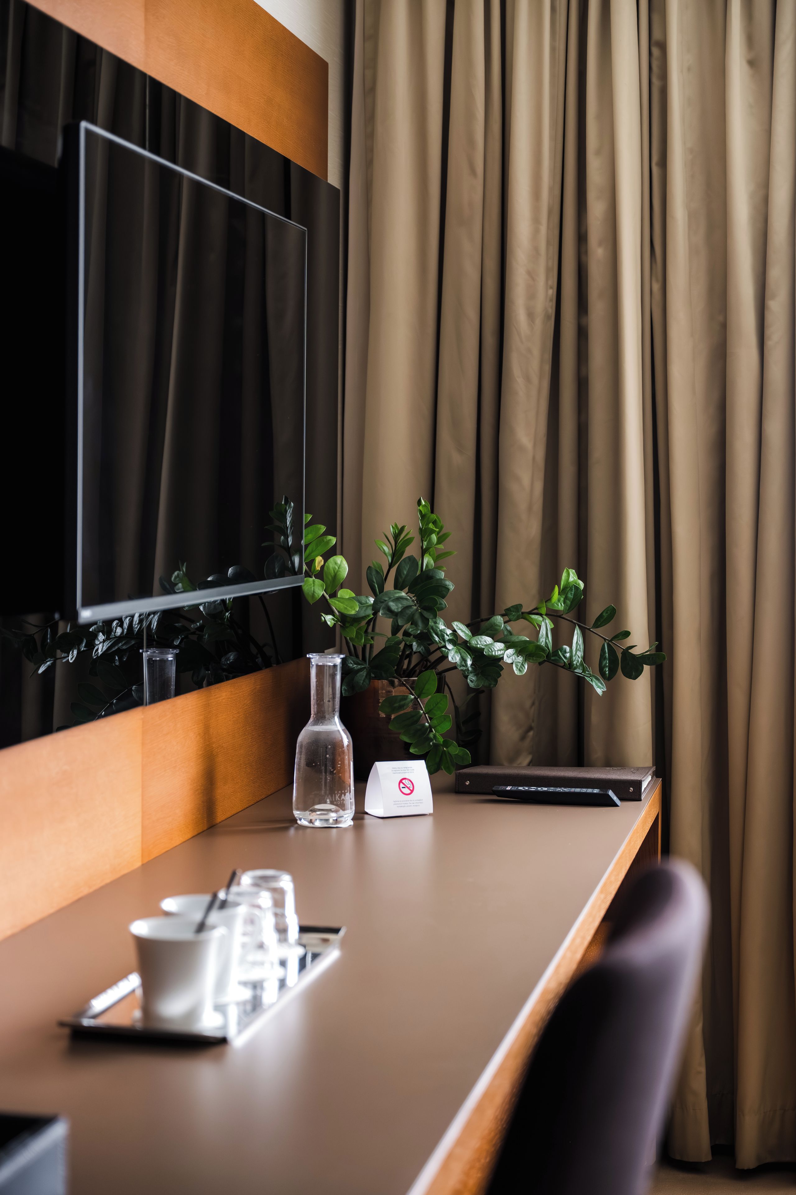 A hotel room with a flat-screen TV mounted on the wall, a wooden desk with a plant, a glass of water, a remote control, and a small white device.