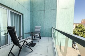 A balcony with a table and two chairs, a window with curtains, and a view of a parked car and buildings. - Mikado Hotel (Nitra)