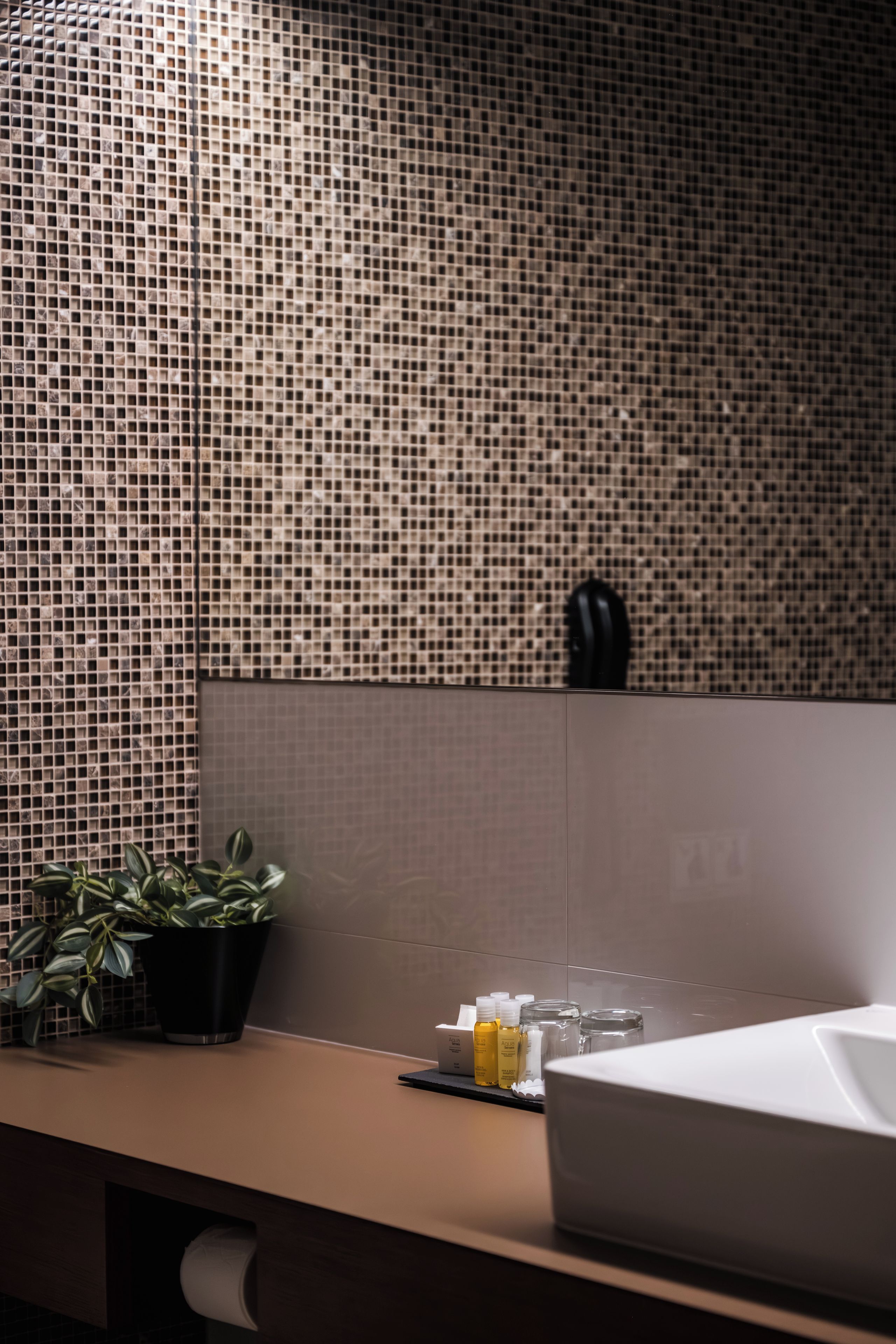 Un baño con salpicadero de azulejos, un lavabo, un espejo, una planta en maceta y artículos de aseo en la encimera.