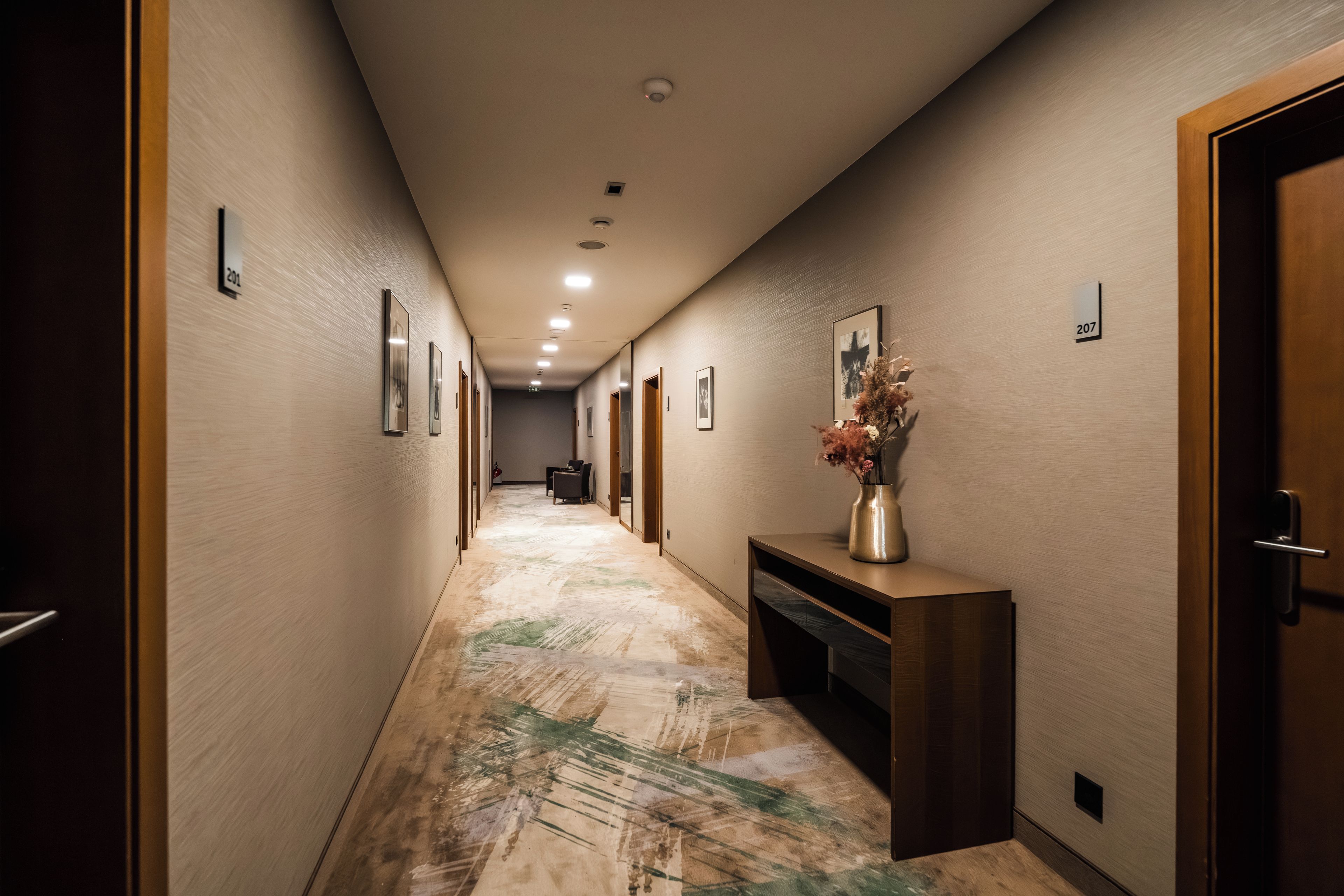 A hotel hallway with framed artwork on the walls, a reception desk with a vase of flowers, and numbered doors.