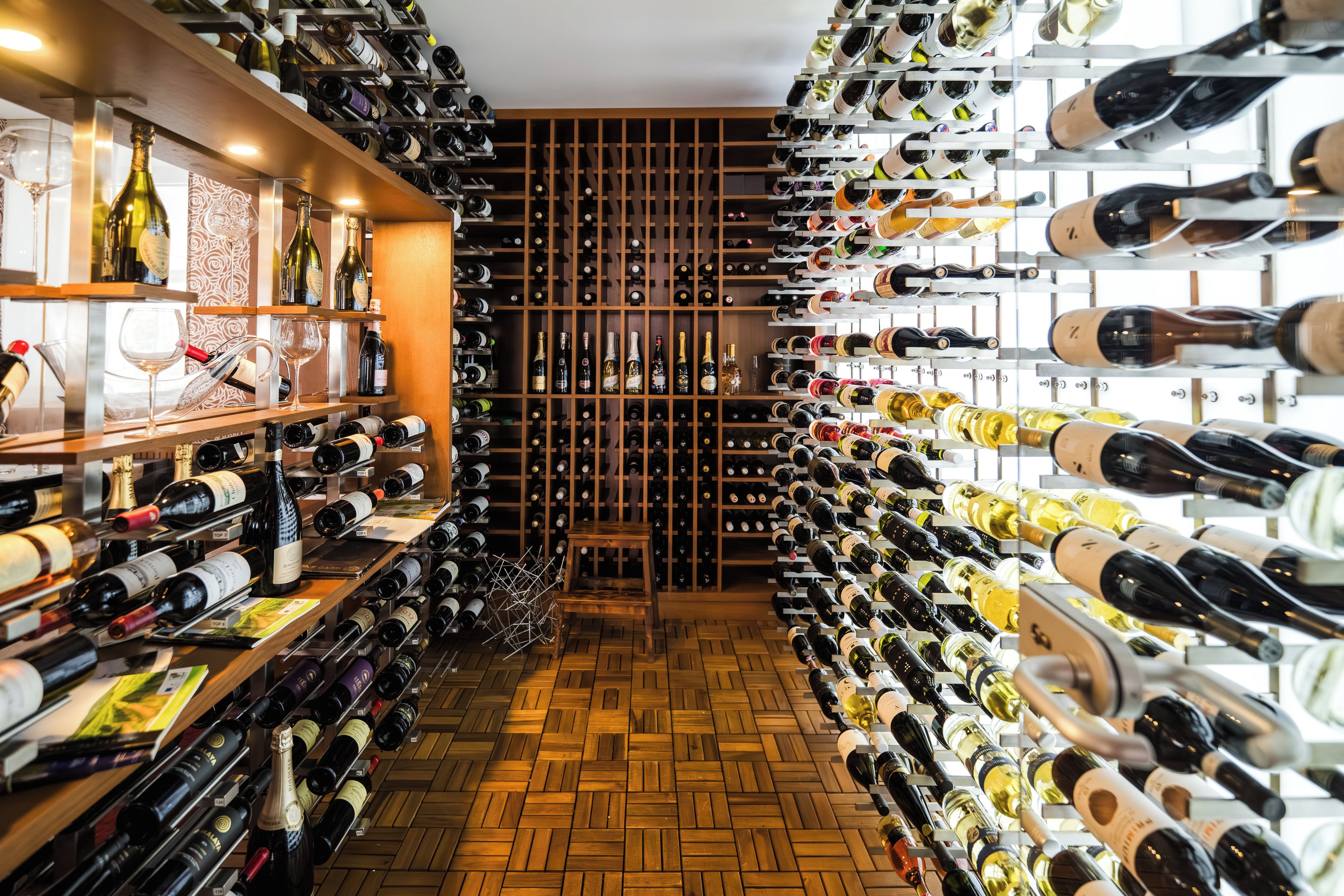 A wine cellar with rows of wine bottles on shelves and racks, wooden flooring, and a central aisle.
