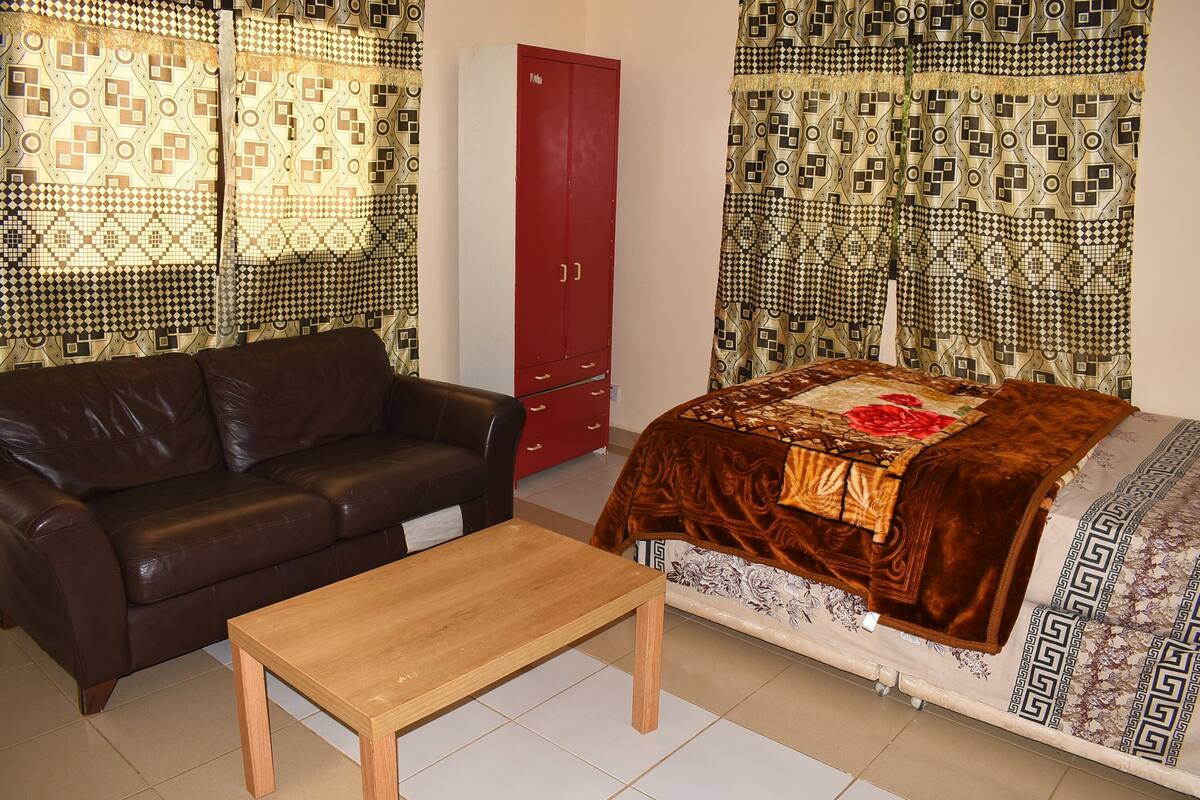 A room with a brown leather sofa, a wooden table, a red wardrobe, and a bed with patterned bedding.
