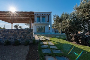 A modern two-story house with a stone wall, a wooden pergola, and a stone pathway leading to the entrance.