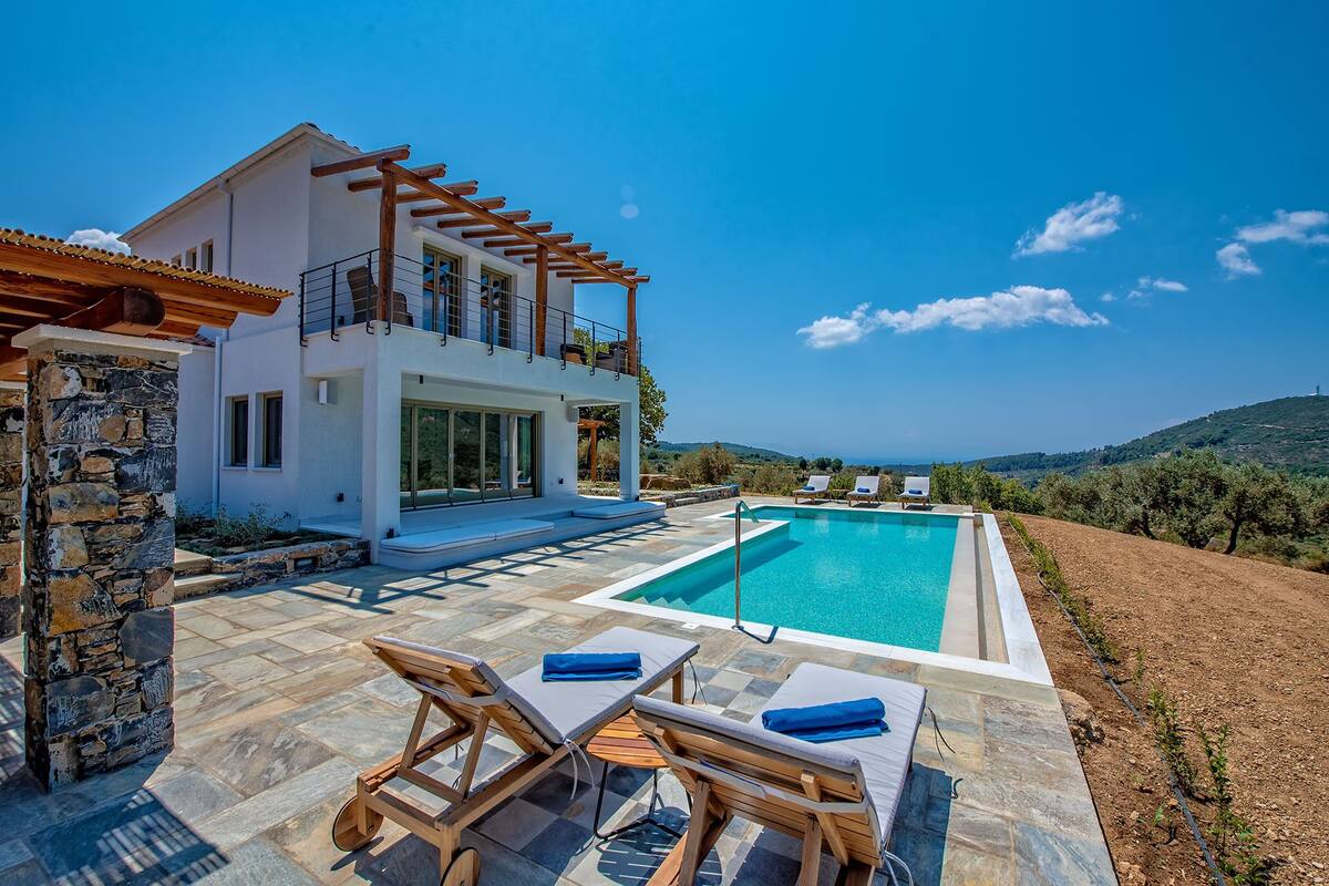 A modern villa with a pool, stone pillars, and a view of the hills.