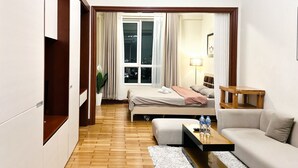 Moderni hotellihuone, jossa on puulattia, sängynpääty, sohva, pieni pöytä, jossa on vesipulloja, ja suuri ikkuna, jossa on verhot.