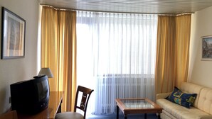 A hotel room with a television on a wooden table, a chair, a glass-top coffee table, a sofa, and a large window with curtains. - Hotel Lust (Hoechst im Odenwald)