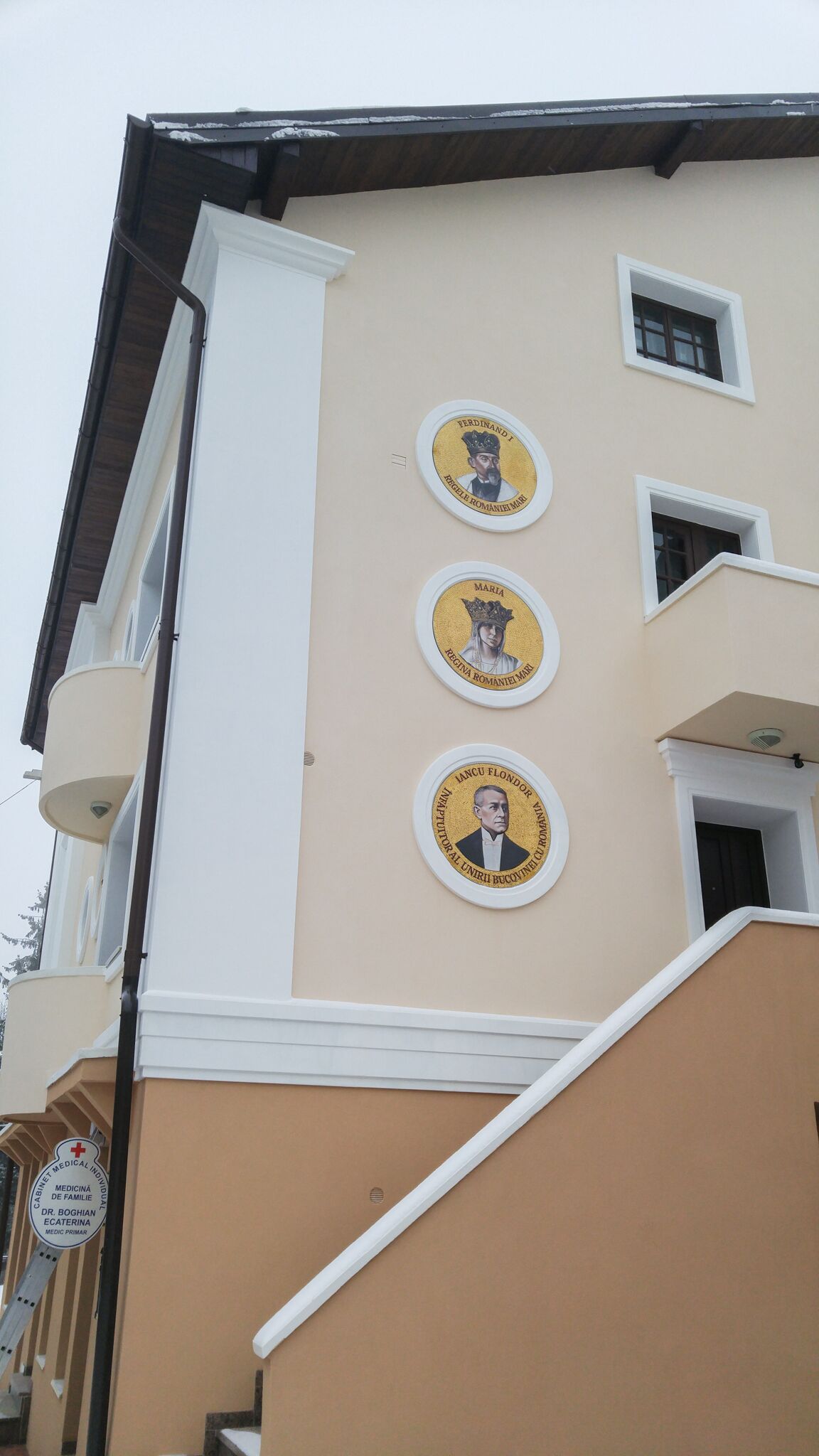 A building with a beige facade, a white staircase, and three circular plaques with portraits and names.