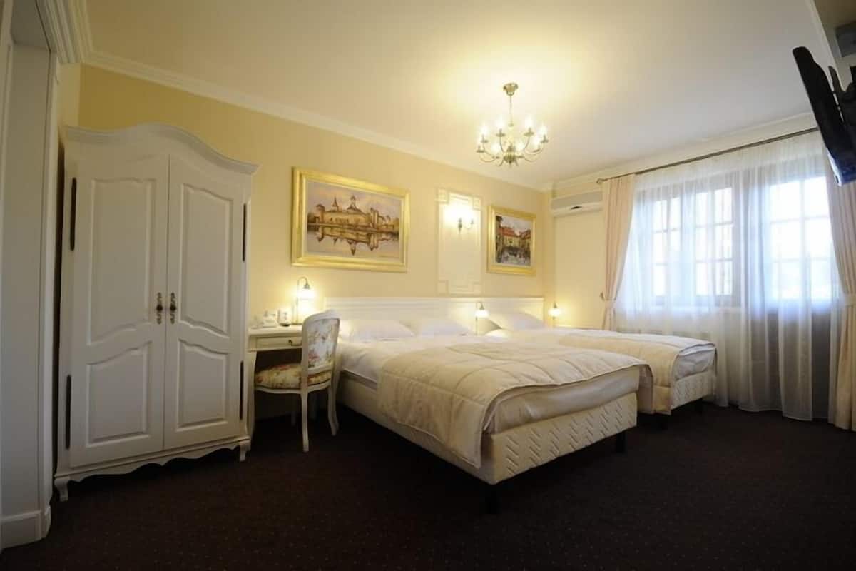 Habitación de hotel con una cama grande, un escritorio y una silla. Hay dos cuadros en la pared, una araña y un amplio ventanal con cortinas.