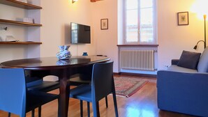A cozy living room with a wooden ceiling, a dining table with chairs, a TV, a radiator, and a window with curtains.