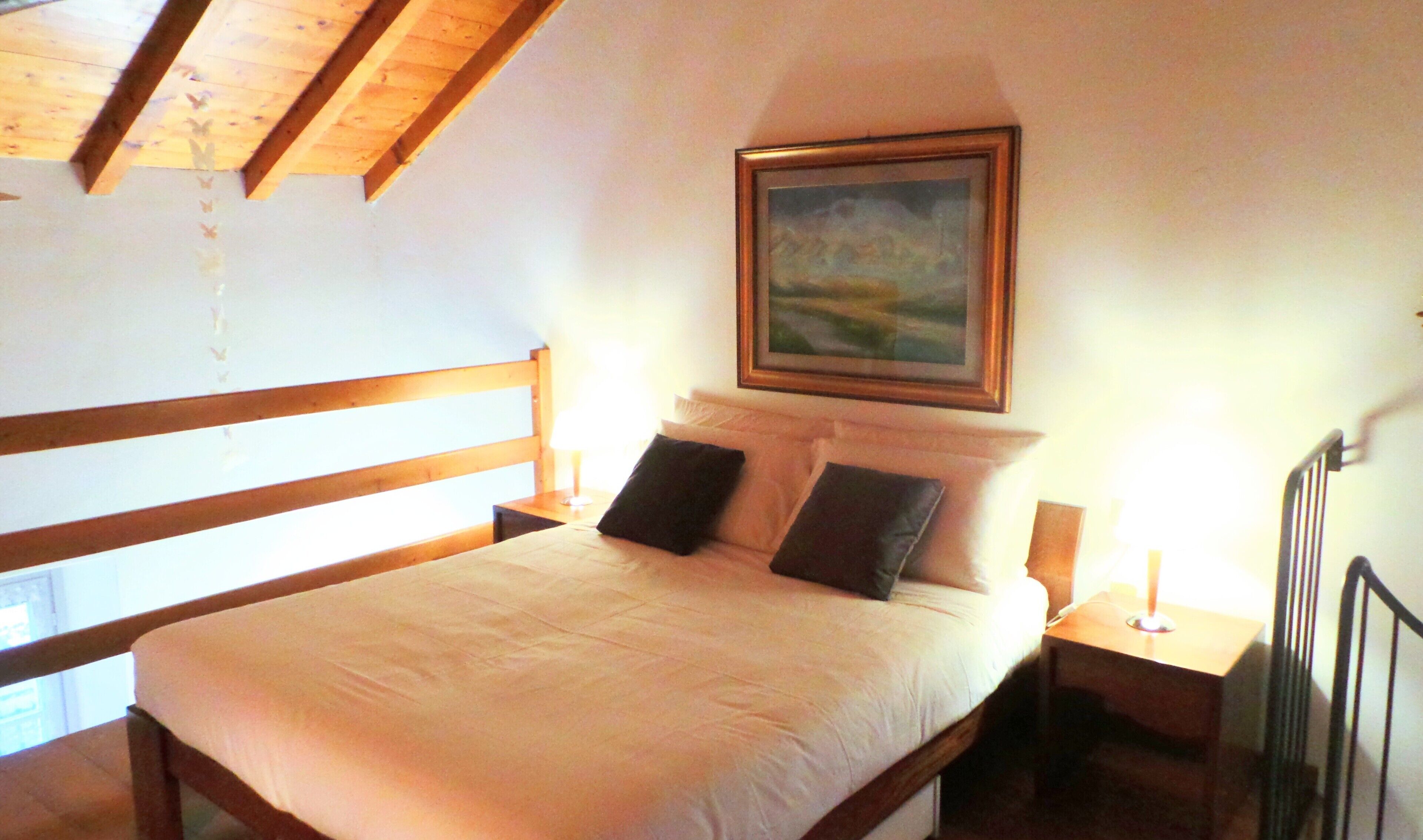 A bedroom with a wooden headboard, a bed with white bedding, two pillows, a nightstand with a lamp, and a framed painting on the wall.