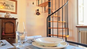 A dining table with plates, cutlery, and a glass of water. A wooden cabinet and a framed picture are on the wall. A spiral staircase with wooden steps and black railings is visible.