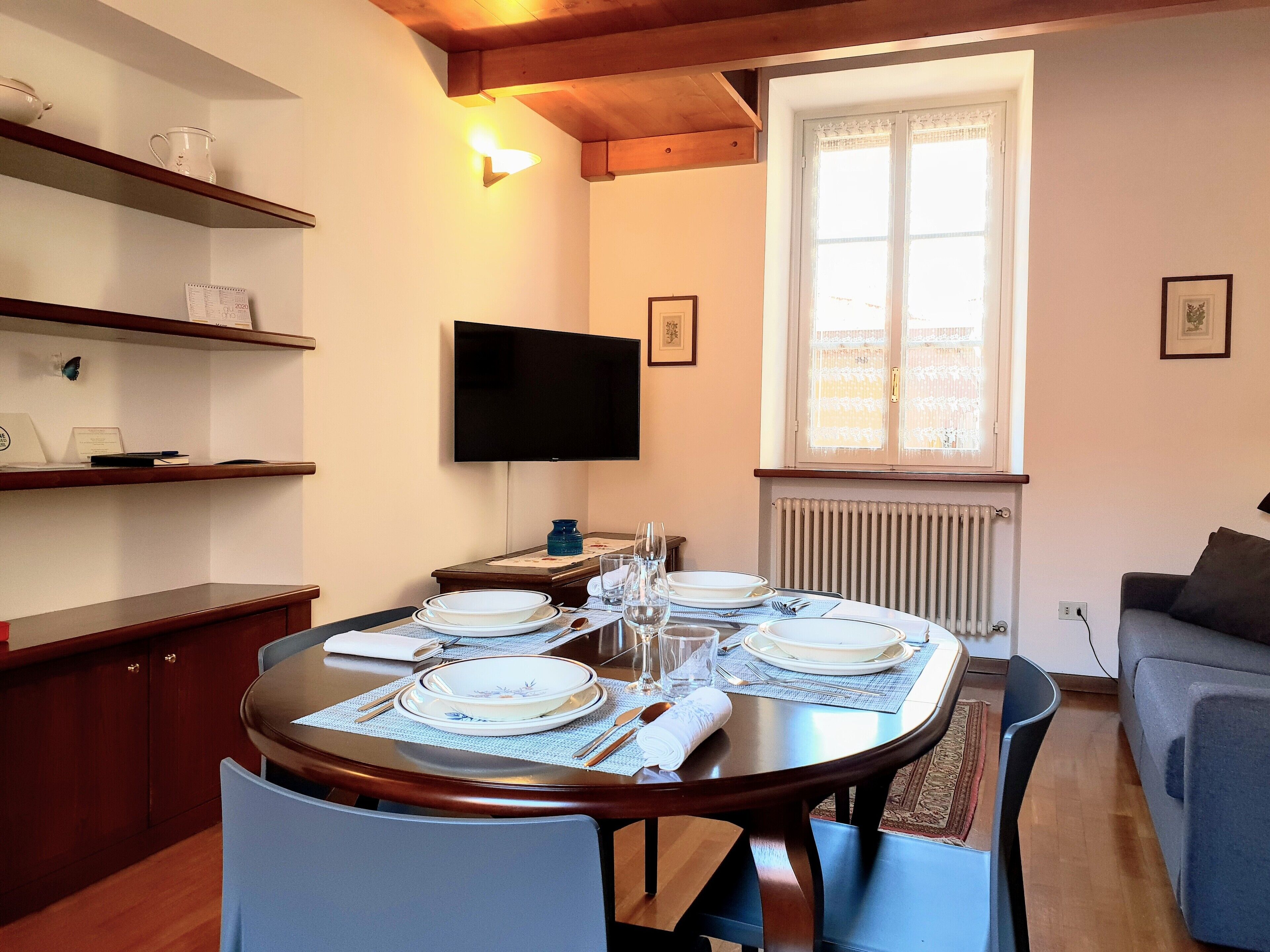 A dining area with a round table set for four, a television mounted on the wall, and a window with a view of the outside.